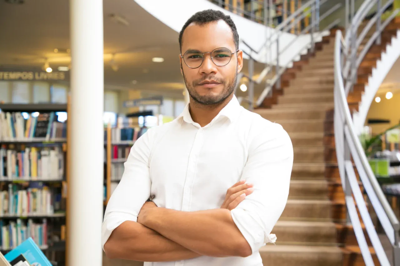 Homem em uma biblioteca com braços cruzados, simbolizando a seriedade e o foco necessários em um curso de inglês para iniciante.